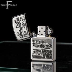 May include: A silver-colored lighter with an open lid, featuring engraved car designs. The lighter's body displays a car, its rear, and a steering wheel. The lid has Japanese text and graphic designs. The brand name "Flint & Forge" is at the top.