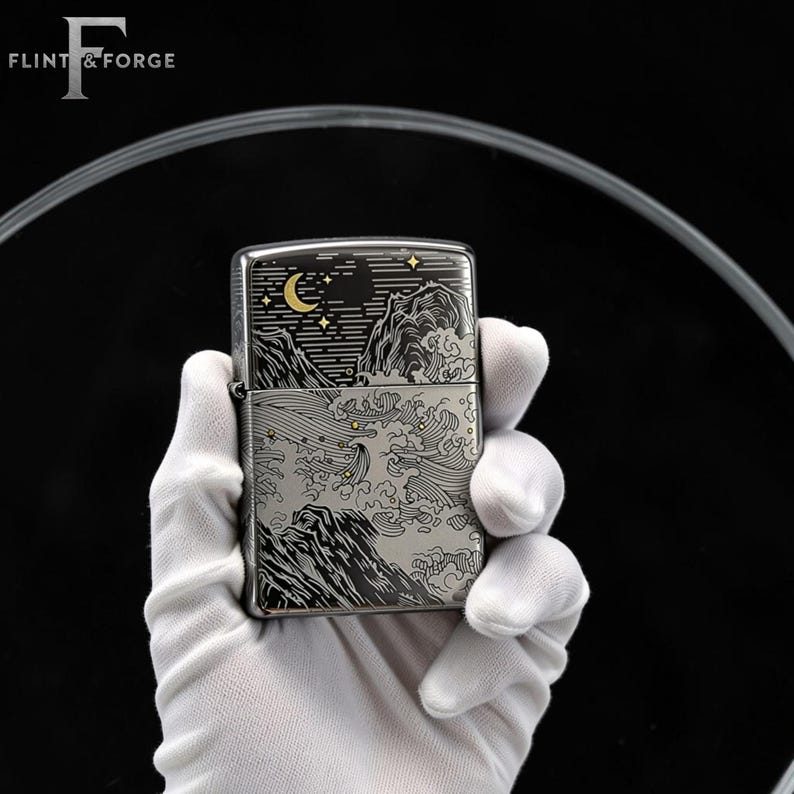 May include: A silver metal lighter with a detailed engraved design. The design features a mountain landscape with a crescent moon, stars, and stylized waves. The lighter is held by a gloved hand, with the brand name "Flint & Forge" visible.