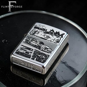 May include: A silver Zippo lighter with a black graphic design featuring race cars and Japanese text. The lighter has the text "SMACK!" and "ALWAYS READY" on it. The bottom of the lighter reads "BRADFORD, PA. MADE IN U.S.A."