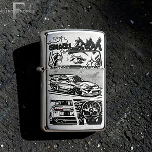 May include: A silver metal lighter with a detailed graphic design. The design includes Japanese text, a car, and the words "ALWAYS READY TO STREET RACE". The lighter is rectangular and has a hinged lid.
