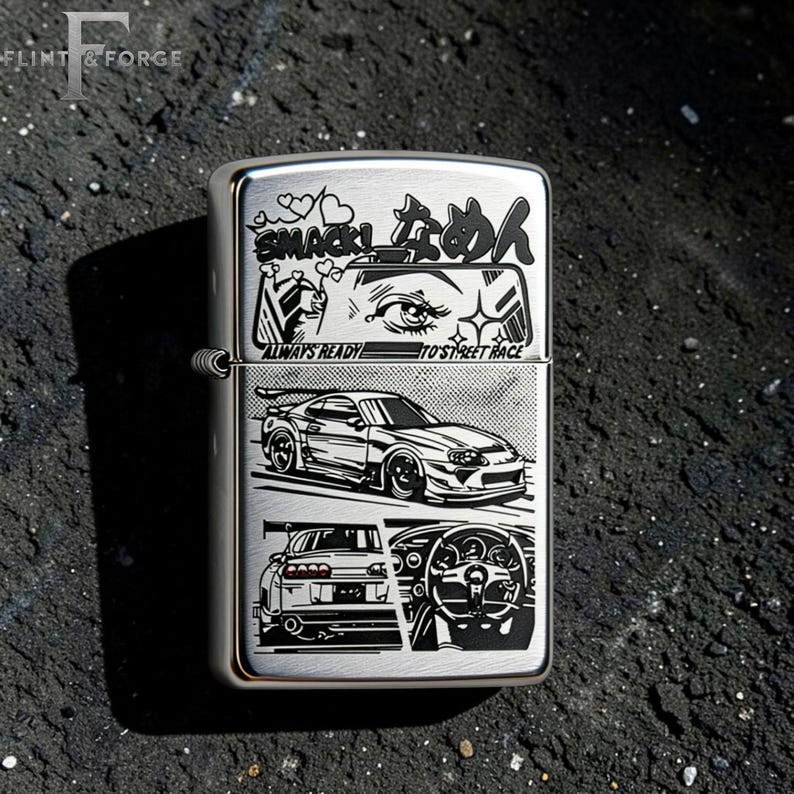 May include: A silver metal lighter with a detailed graphic design. The design includes Japanese text, a car, and the words "ALWAYS READY TO STREET RACE". The lighter is rectangular and has a hinged lid.