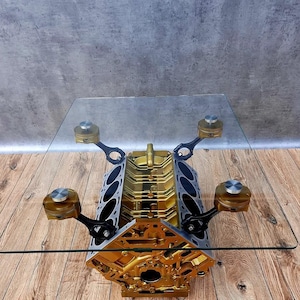 May include: A unique coffee table featuring a gold-colored engine block base with a glass top. The engine block has silver and black accents, with pistons attached. The table sits on a wooden floor.