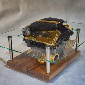 May include: A distinctive coffee table with a car engine, highlighted with gold detailing, under a clear glass surface. The engine sits on a wooden base, supported by silver legs. The table measures approximately 122 cm long, 76 cm wide, and 46 cm high.