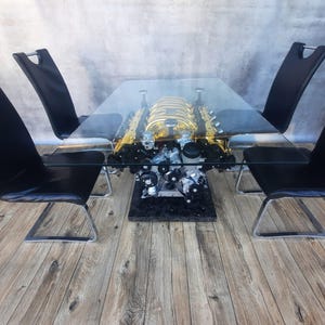 May include: A rectangular glass table with a car engine base and four black chairs. The engine is gold and black, and the chairs have chrome legs. The table is set on a wooden floor.