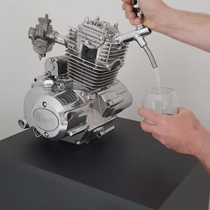 Może przedstawiać: Chromowany silnik motocyklowy, zmodyfikowany kranem, dozujący płyn do szklanki. Silnik zamontowany jest na czarnym cokole. Silnik ma napis "ROMET".