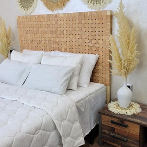 Op de afbeelding: Een slaapkamer met een geweven houten hoofdbord, wit beddengoed en wanddecoraties. Een houten nachtkastje met een witte vaas en gedroogde bloemen staat naast het bed. De kamer heeft een warme, uitnodigende uitstraling.