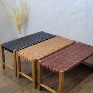 Op de afbeelding: Drie houten banken met geweven leren zittingen in zwart, bruin en beige. De banken hebben een rechthoekige vorm en worden ondersteund door vier houten poten. De achtergrond toont wanddecoraties en een vaas met gedroogde planten.