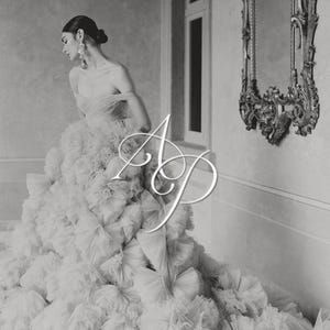 May include: A floor-length off-the-shoulder gown featuring a voluminous skirt constructed from layers of ruffled tulle fabric. The bodice has a sweetheart neckline and draped sleeves. The letters AP are overlaid in white in the center of the image.