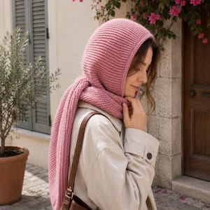 May include: A knitted pink hood and scarf set. The hood covers the head and neck, with a scarf that hangs down the front. The set is made of a soft, textured yarn. The person is wearing a light-coloured coat and carrying a brown leather bag.