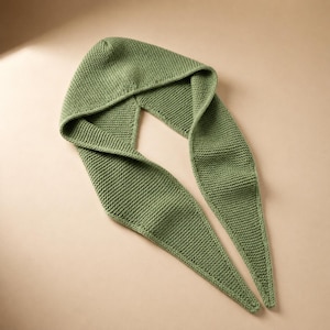 May include: A knitted green hood with long, pointed scarf ends. The hood and scarf are made of a textured knit fabric. The scarf is laying on a neutral background.