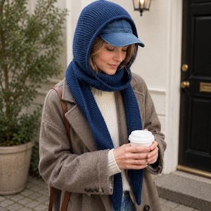 Puede incluir: Una mujer con un conjunto de capucha y bufanda de punto azul marino sobre una gorra de béisbol vaquera. También lleva un abrigo marrón, un suéter blanco y vaqueros azules. Sostiene una taza de café de papel blanca.