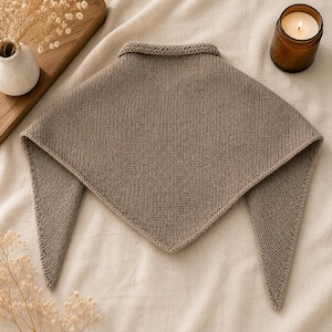 Puede incluir: Un chal de punto de color topo con cuello y dos extremos puntiagudos. El chal está hecho de un hilo suave y texturizado. El fondo incluye una tabla de cortar de madera, un pequeño jarrón blanco y una vela encendida en un tarro de cristal.
