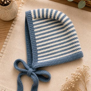Puede incluir: Un gorro de punto con un patrón de rayas horizontales azules y blancas. El gorro tiene un borde acanalado de color azul sólido a lo largo de la abertura facial e incluye dos lazos de punto largos y a juego de color azul anudados en la barbilla.
