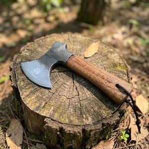 May include: A hand-forged steel hatchet with a dark, textured metal head featuring an engraved Viking-style emblem and the name David. The wooden handle is inscribed with the text Stand strong, strike true and includes a black lanyard.