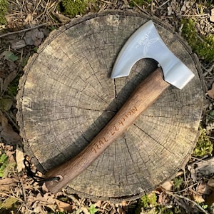 May include: A silver axe with a wooden handle, engraved with the name "David" and the date "March 27 2022". The axe is resting on a weathered tree stump, showcasing the wood grain.