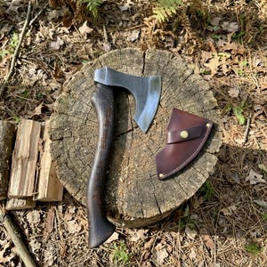 Puede incluir: Un hacha de mano con mango de madera oscura con texto grabado y hoja de acero pulido, acompañada de una funda de cuero marrón con cierre de presión de latón. La cabeza del hacha tiene un filo curvo y un acabado metálico liso.