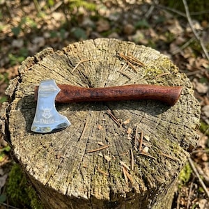 Puede incluir: Un hacha pequeña y decorativa con una cabeza plateada pulida y un mango de madera marrón oscuro. La cabeza del hacha está grabada con un emblema de ciervo y el nombre "David". El mango tiene la inscripción "Strength is not given, it is forged through struggle."