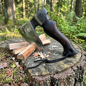 May include: A hand axe with a polished steel blade featuring an engraved Viking helmet icon and the name David. The dark wood handle is engraved with the date April 27, 2026, and includes a black paracord lanyard attached to the base of the grip.