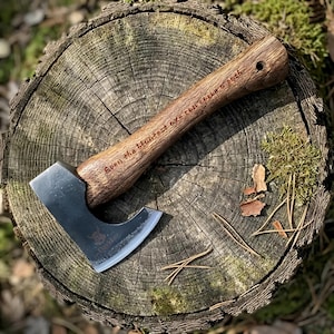 Puede incluir: Un hacha pequeña con una hoja de metal oscuro y un mango de madera. El mango está grabado con el texto "Even the bluntest eye can carve a path". El hacha está sobre un tocón de árbol.