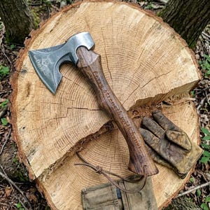 Può includere: Un'accetta in acciaio con manico in legno scuro che presenta l'incisione "Carry strength with honor". La lama in metallo è incisa con un emblema decorativo e il nome "David". Un laccetto in cuoio è fissato al manico dell'accetta.