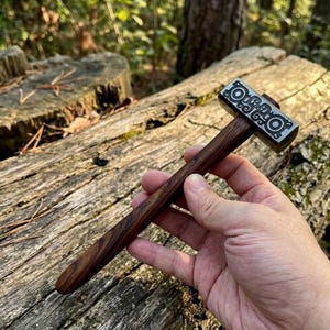 May include: A small hammer featuring a dark brown wooden handle with a visible grain and a rectangular metal head. The metal head is decorated with intricate, symmetrical engraved patterns on its side, showcasing circular and swirling line motifs.