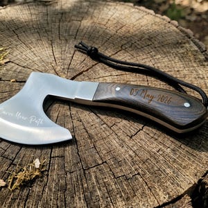 Peut inclure: Petite hache argentée avec un manche en bois foncé, posée sur une souche d'arbre. La lame porte l'inscription "Carve Your Path". Le manche est gravé "05 May 2026". Une corde noire est attachée.