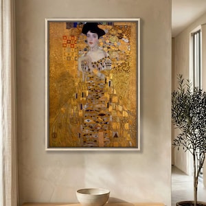 Könnte beinhalten: Ein gerahmter vertikaler Wandkunstdruck mit dem Porträt einer Person mit dunklem Haar, die ein goldfarbenes Kleid mit komplizierten geometrischen Mustern, Augenmotiven und Blattgoldtexturen vor einem reich schimmernden Hintergrund trägt.