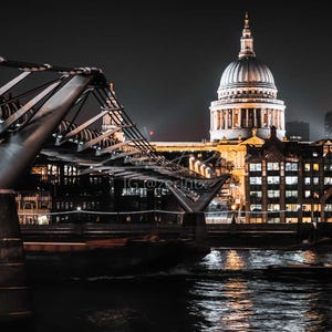 Può includere: Paesaggio notturno di Londra con il Millennium Bridge e la Cattedrale di St Paul. La struttura in acciaio del ponte contrasta con la cupola della cattedrale. Luci della città e una barca sull'acqua illuminano la scena.