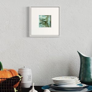 May include: A framed abstract painting with shades of green and blue. The painting is hanging on a white wall above a table with a pitcher, a jar, a basket of fruit, and a stack of plates.