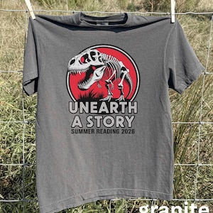 May include: A gray t-shirt with a dinosaur skeleton graphic inside a red circle. The text "UNEARTH A STORY SUMMER READING 2026" is printed below the graphic. The shirt is hanging on a clothesline.