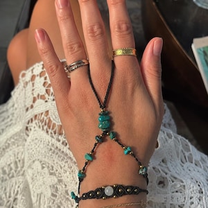 May include: A black cord hand chain featuring turquoise stone beads and small gold-tone accents, connecting a finger loop to a wrist bracelet. The wrist stack includes a black macrame bracelet with a central white stone, a thin gold chain, and a gold-tone link bracelet. Two gold-tone rings are worn on the fingers.