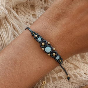 May include: A macrame bracelet featuring dark grey cord woven into a geometric pattern, accented with three light blue round stones and several small gold-toned metal beads. The bracelet has an adjustable cord closure with small beads at the ends.