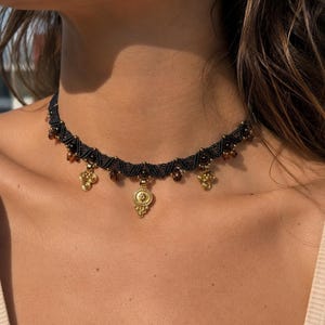 May include: A black macrame choker necklace featuring small dark brown beads woven into the cord. The necklace is adorned with several gold-toned metal charms, including a central circular pendant and smaller triangular accents hanging along the band.