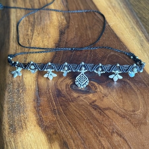 May include: A black macrame choker necklace featuring a central diamond-shaped silver-tone metal charm, accented with smaller silver-tone metal flower-shaped charms and tiny beads woven into the dark cord, finished with adjustable thin black ties.