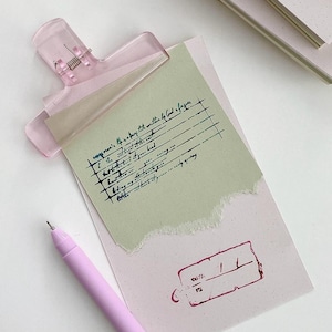 May include: A light pink translucent hair clip secures a piece of sage green paper with handwritten text and a torn edge onto a pale pink rectangular card. The card features a dark red stamped date box and is accompanied by a light pink ballpoint pen.