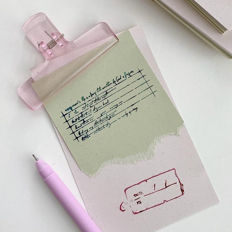 May include: A light pink translucent hair clip secures a piece of sage green paper with handwritten text and a torn edge onto a pale pink rectangular card. The card features a dark red stamped date box and is accompanied by a light pink ballpoint pen.