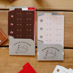 Puede incluir: Dos juegos de pegatinas de índice en marrón oscuro y beige claro, cada uno con tres agujeros para archivador y pestañas numeradas del 01 al 11. El empaque es una tarjeta gris con el texto Index Sticker with loose-leaf y Benstore Design.