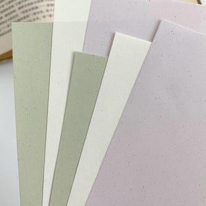 May include: A collection of rectangular paper sheets in shades of sage green, off-white, and soft lavender. Each sheet features a distinct speckled texture with small dark flecks distributed across the surface, arranged in a fanned-out stack.