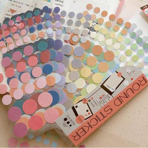 May include: A collection of small, circular adhesive labels in various pastel and vibrant colors, including pink, blue, yellow, and green. The stickers feature a textured surface and are presented with a branded packaging card labeled ROUND STICKER.