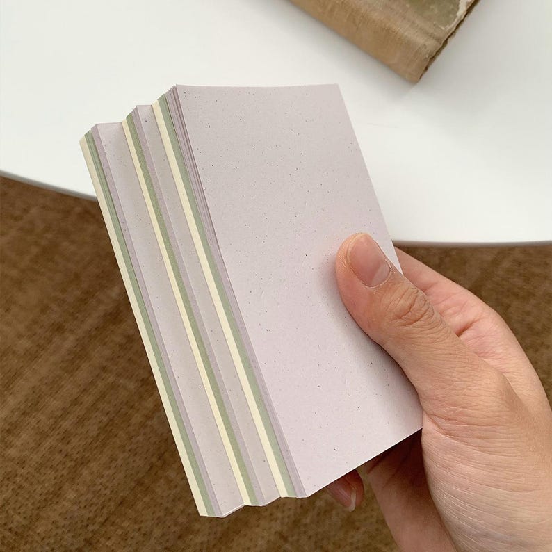 May include: A stack of rectangular paper cards with a speckled texture. The cards feature a layered color scheme consisting of light lavender, pale green, and cream white edges, creating a distinct striped effect along the side of the stack.