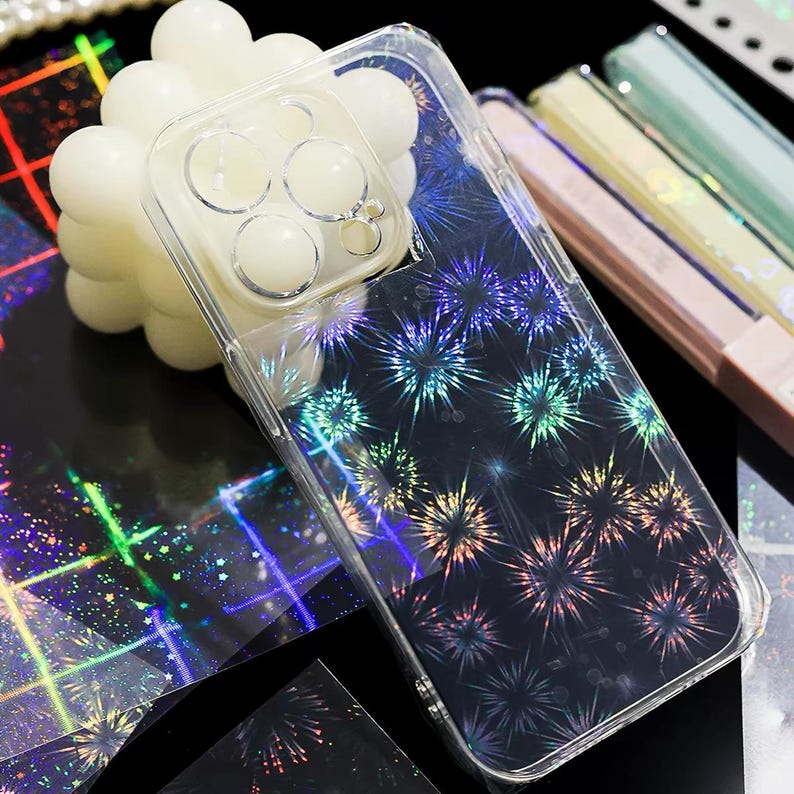 May include: A clear smartphone case featuring a holographic firework pattern that reflects light in various colors. The case has a dedicated cutout section for a multi-lens camera array, showcasing a transparent design that allows the device color to show through.