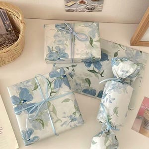 May include: Three gifts wrapped in white paper featuring a blue floral pattern with green leaves. Two rectangular boxes are tied with light blue ribbon, and one cylindrical gift is wrapped like a candy with blue ribbon tied at both ends.