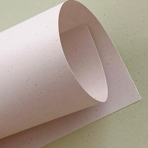 May include: A single sheet of light pink paper with a speckled texture, rolled into a curved shape. The paper features small, dark, randomly distributed flecks across its surface, creating a subtle, natural, and fibrous appearance throughout.
