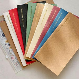 May include: A collection of slim, rectangular notebooks arranged in a fan shape. Each notebook cover features the text TRAVEL NOTES and Benstore Design in gold foil. The covers come in white, red, black, green, tan, cream, maroon, and blue.