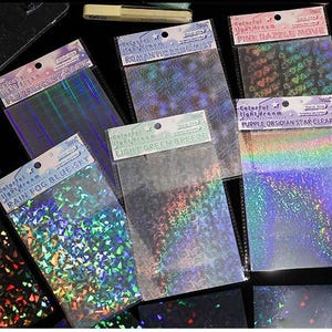 May include: A collection of rectangular sheets of holographic flash film in various patterns and colors. Each sheet is packaged in a clear plastic sleeve with a header card featuring text such as Fluorescent Magic, Romantic Blue Mist, Pink Dazzle Move, Rain Fog Blue Sky, Light Green Breeze, and Purple Obsidian Star Clear.