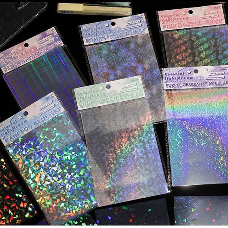 May include: A collection of rectangular sheets of holographic flash film in various patterns and colors. Each sheet is packaged in a clear plastic sleeve with a header card featuring text such as Fluorescent Magic, Romantic Blue Mist, Pink Dazzle Move, Rain Fog Blue Sky, Light Green Breeze, and Purple Obsidian Star Clear.