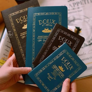 May include: Four rectangular notebooks in dark brown and teal colors, featuring gold foil text that reads DOUX ET AÉRIEN, SOMETIMES SOMETIMES, and a small cat illustration. Each notebook is decorated with a gold border containing small utensil icons.