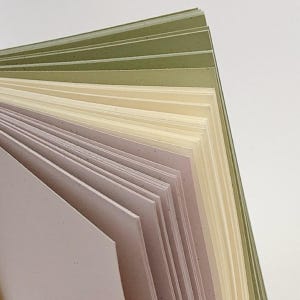 May include: A stack of rectangular paper sheets fanned out to display a gradient of colors. The sheets transition from a soft dusty pink at the front to a creamy off-white in the middle, ending with shades of sage green at the back edge.