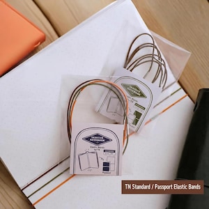 Könnte beinhalten: Zwei Sets Traveler Notebook Gummibänder in transparenter Kunststoffverpackung. Jedes Set enthält dünne, kreisförmige Gummikordeln in Braun, Orange und Grün sowie ein Papieretikett mit dem Text Traveler Notebook und Abbildungen von Notizbüchern.