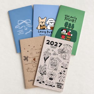 May include: Five rectangular notebooks with varied cover designs. One blue notebook features a white ribbon graphic and text. A light blue notebook shows two cartoon dogs. A green notebook displays a character with text "the daily YOGURT". Two tan notebooks feature line art illustrations, birds, and "2027" text.